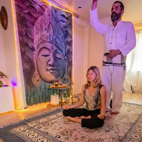 Ali standing with a raised hand over a woman seated in meditation in a candlelit healing room