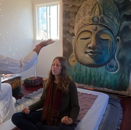 Woman seated in meditation on a treatment bed beside a carved Buddha face painting