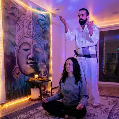 Ali with arms raised over a woman meditating in a violet-lit room